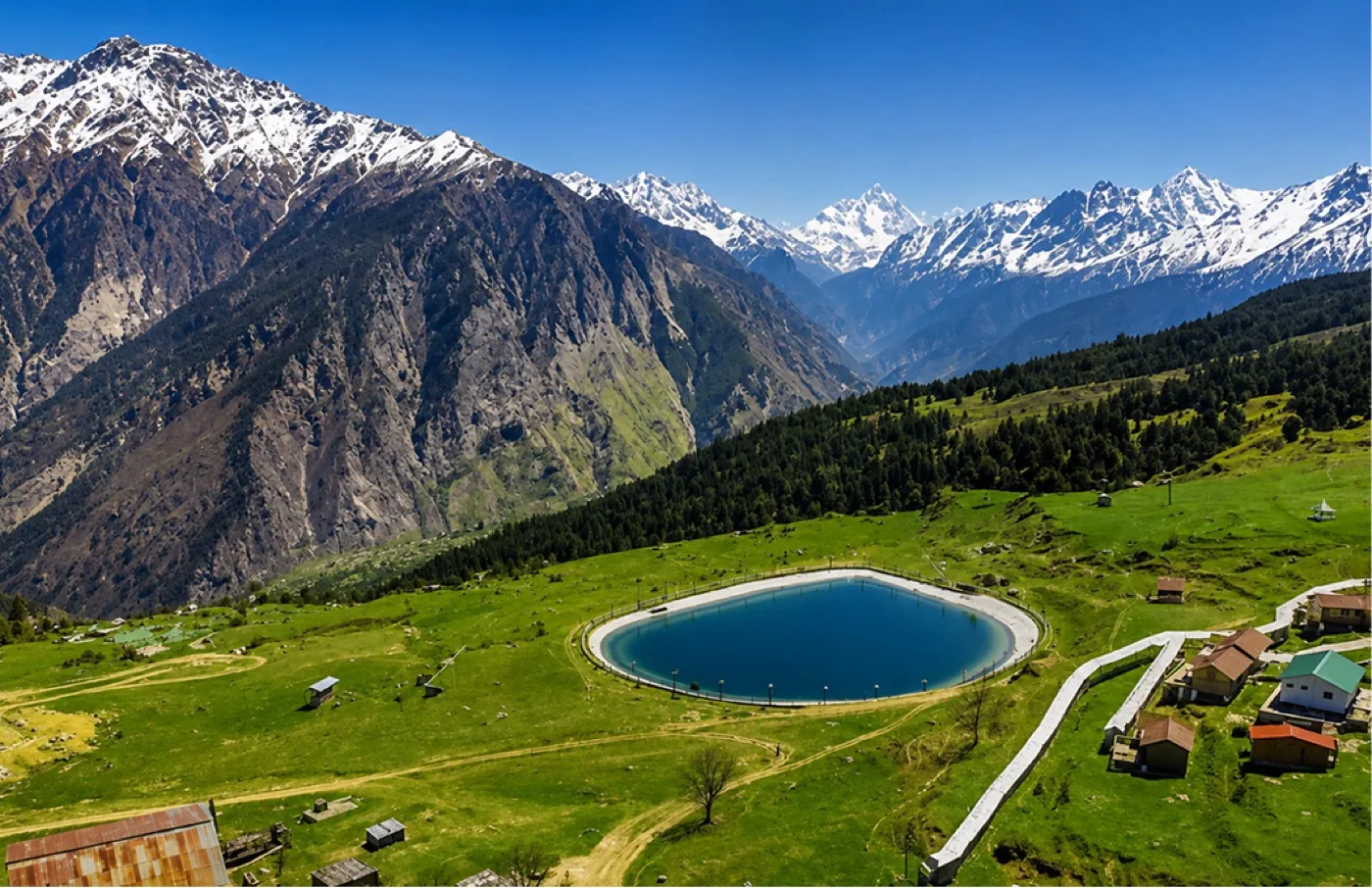 Trekkers crossing a green meadow below a snow-covered Himalayan range