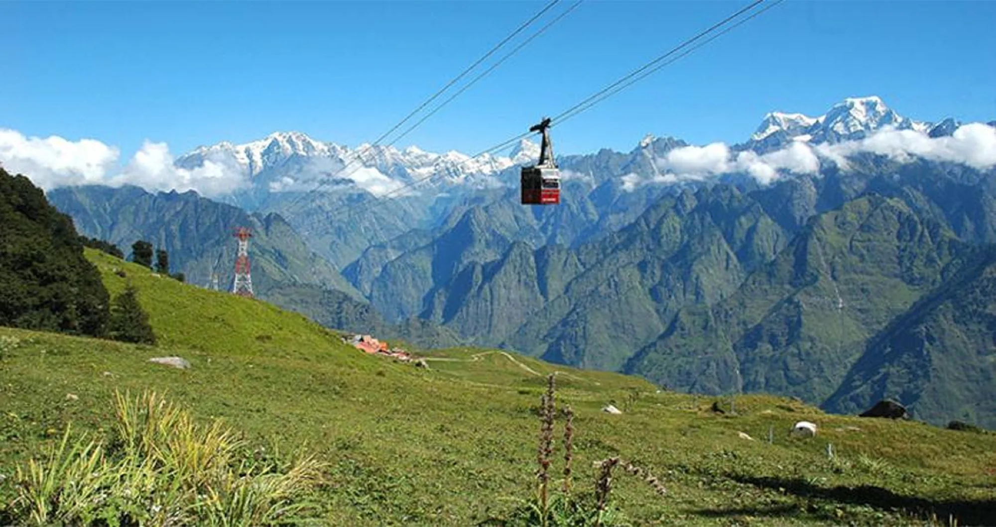 A cable car crossing a green summer meadow, with a snow-capped range standing beyond the valley