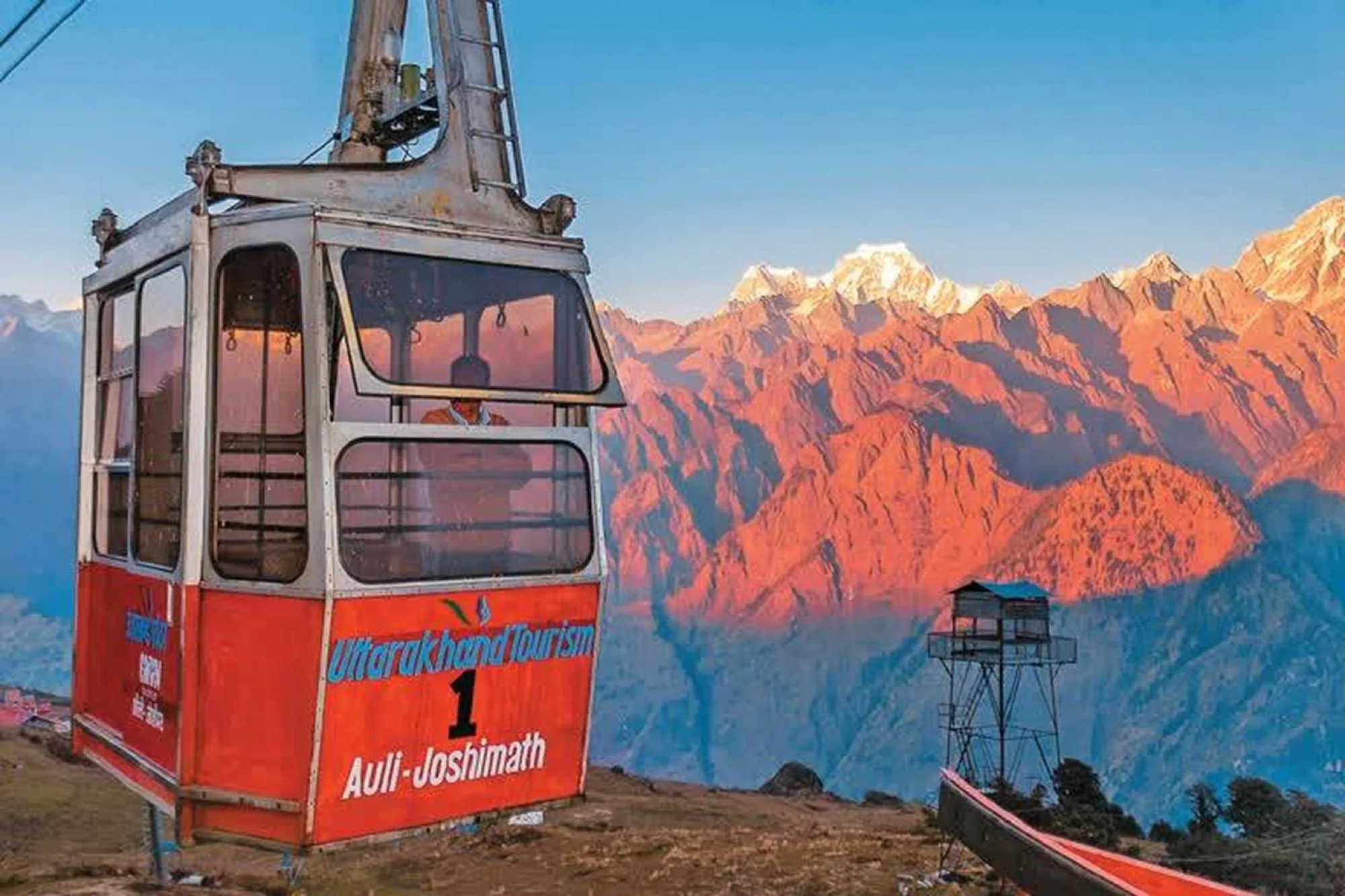 A red cable car cabin lettered Auli-Joshimath at a top station, with sunset light burning across the snow peaks behind it