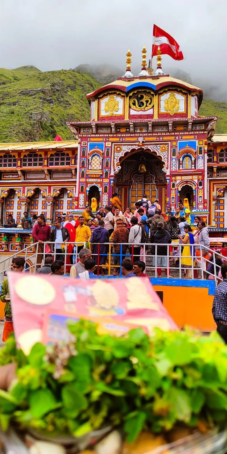 A crowd on the steps and forecourt of a brightly painted temple, an offering of sweets and tulsi leaves held up in the foreground, cloud on the green ridge behind