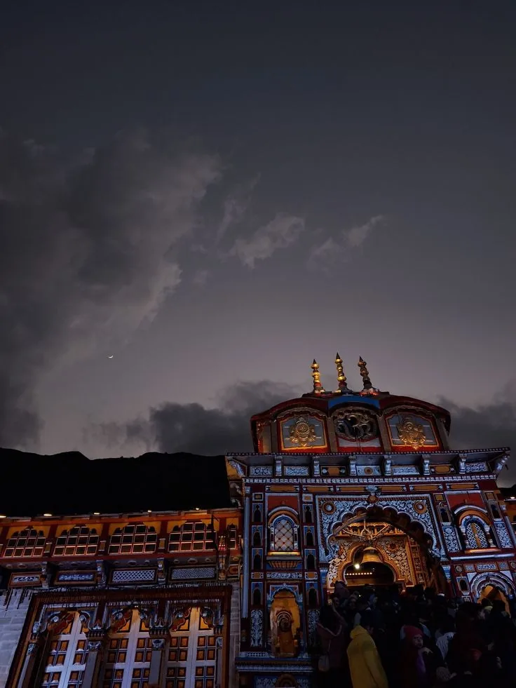 A brightly painted temple facade lit against a near-black sky with a thin crescent moon, the ridge behind in silhouette