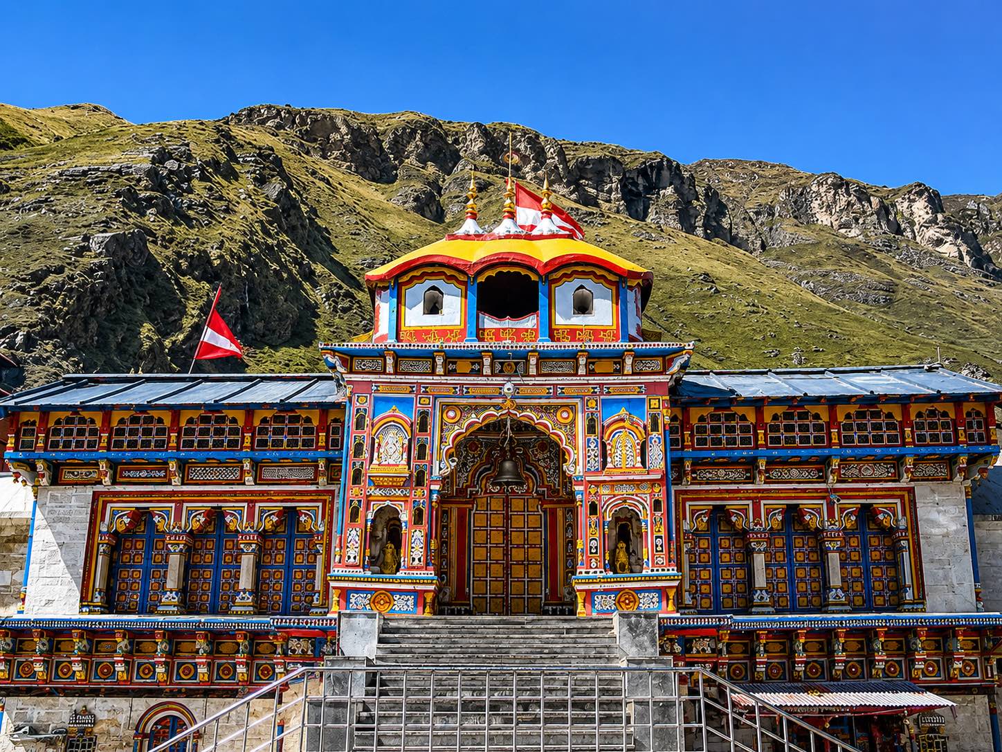 A tall temple facade painted in blue, red and yellow with a carved arched doorway and a tiered gold-finialled roof, a steep treeless slope of grass and rock rising close behind it