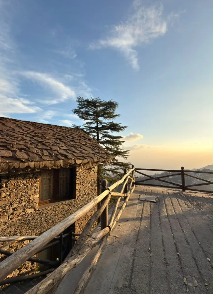 A slate-roofed stone cottage and a lone deodar above Mussoorie at sunset, the hillside town below