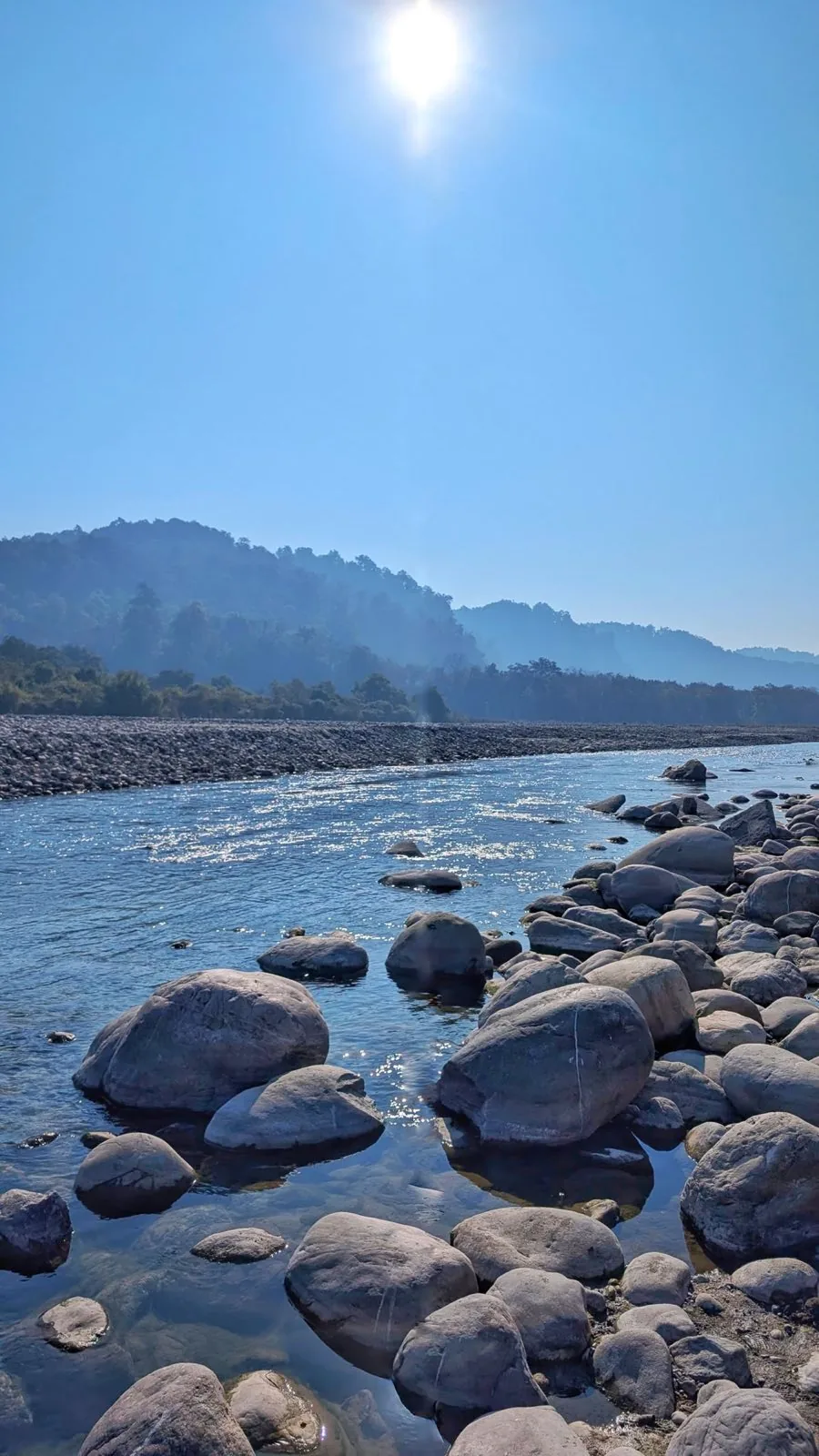 A shallow river running between rounded grey boulders, with wooded ridges hazy behind it under a high sun