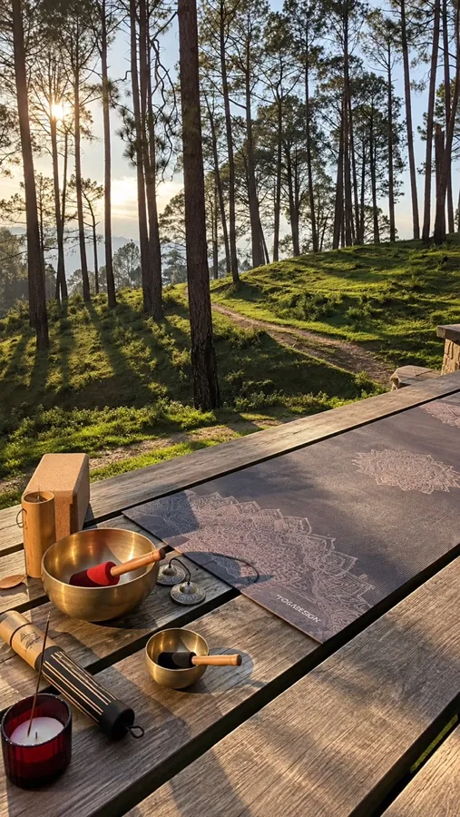 A yoga mat and singing bowls on a wooden deck in a pine forest at sunset
