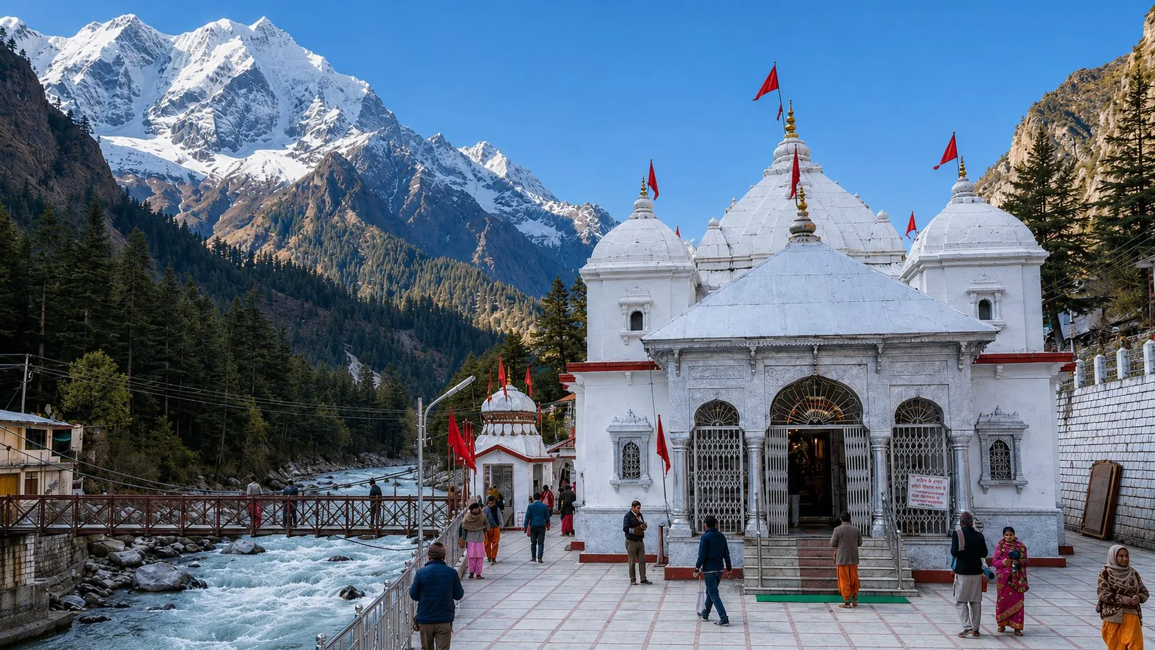 A white multi-shikhara temple complex on a paved riverside terrace, a footbridge over the torrent and a snow wall closing the valley behind