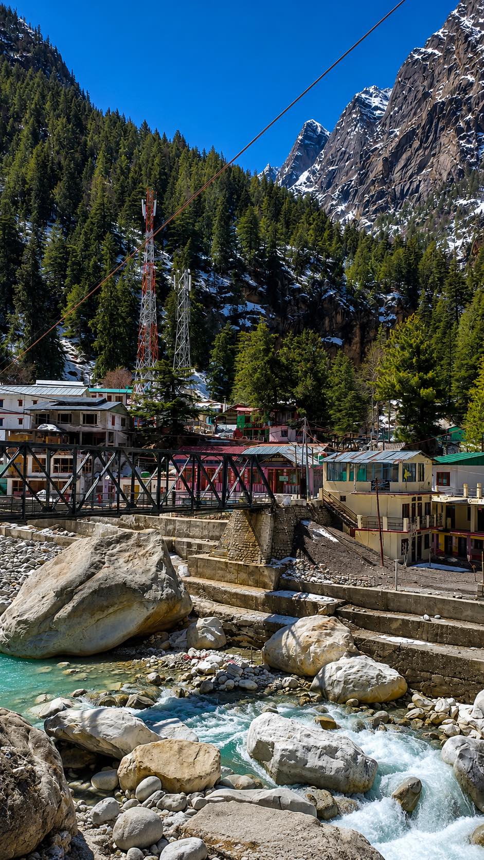 A steel truss bridge over a turquoise boulder river, painted tin-roofed houses along the far bank below deodar forest and snow-streaked crags