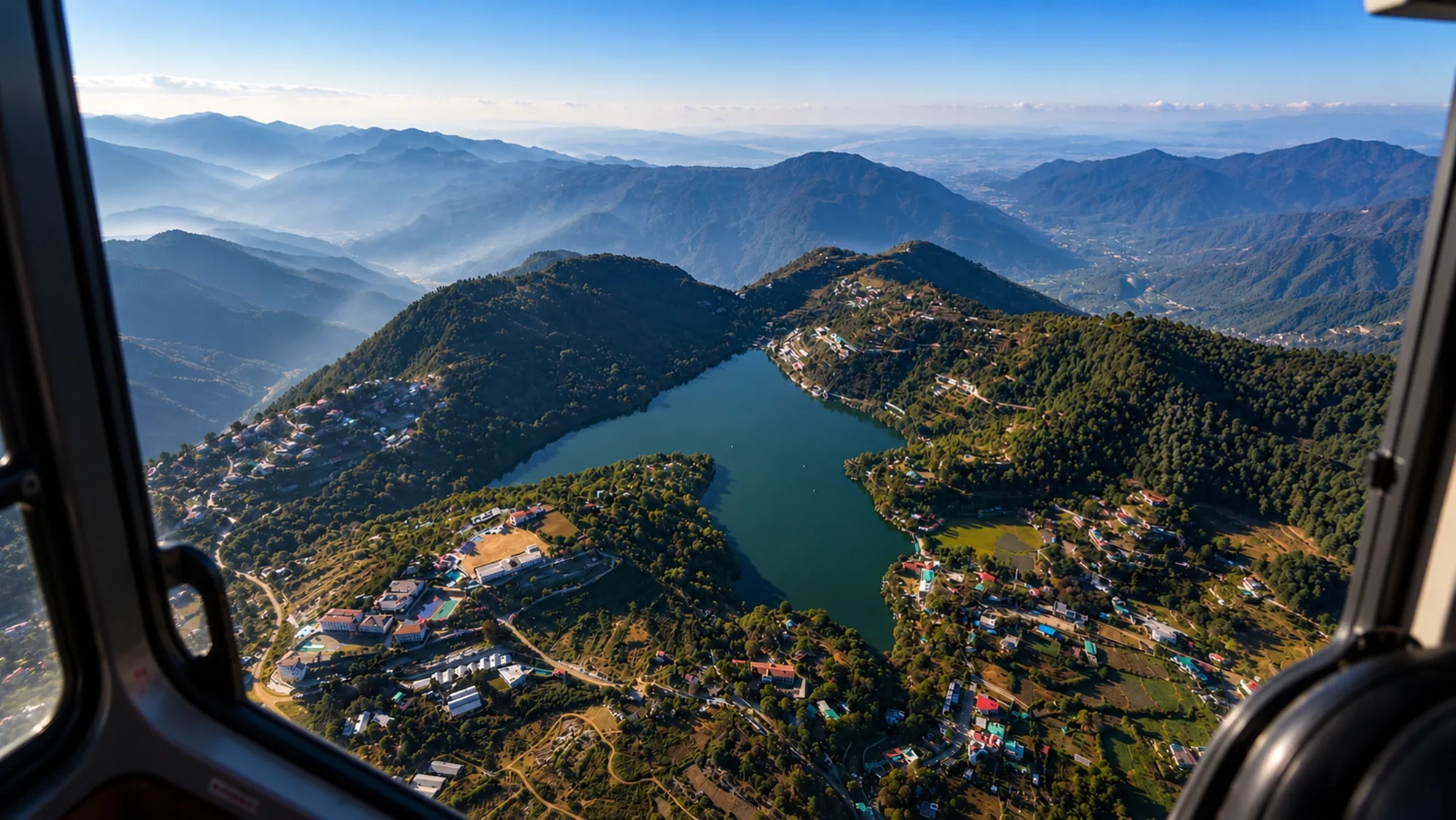 An aerial view through a helicopter door of a Himalayan lake held between forested ridges, hillside towns along both shores and ranges fading to the horizon