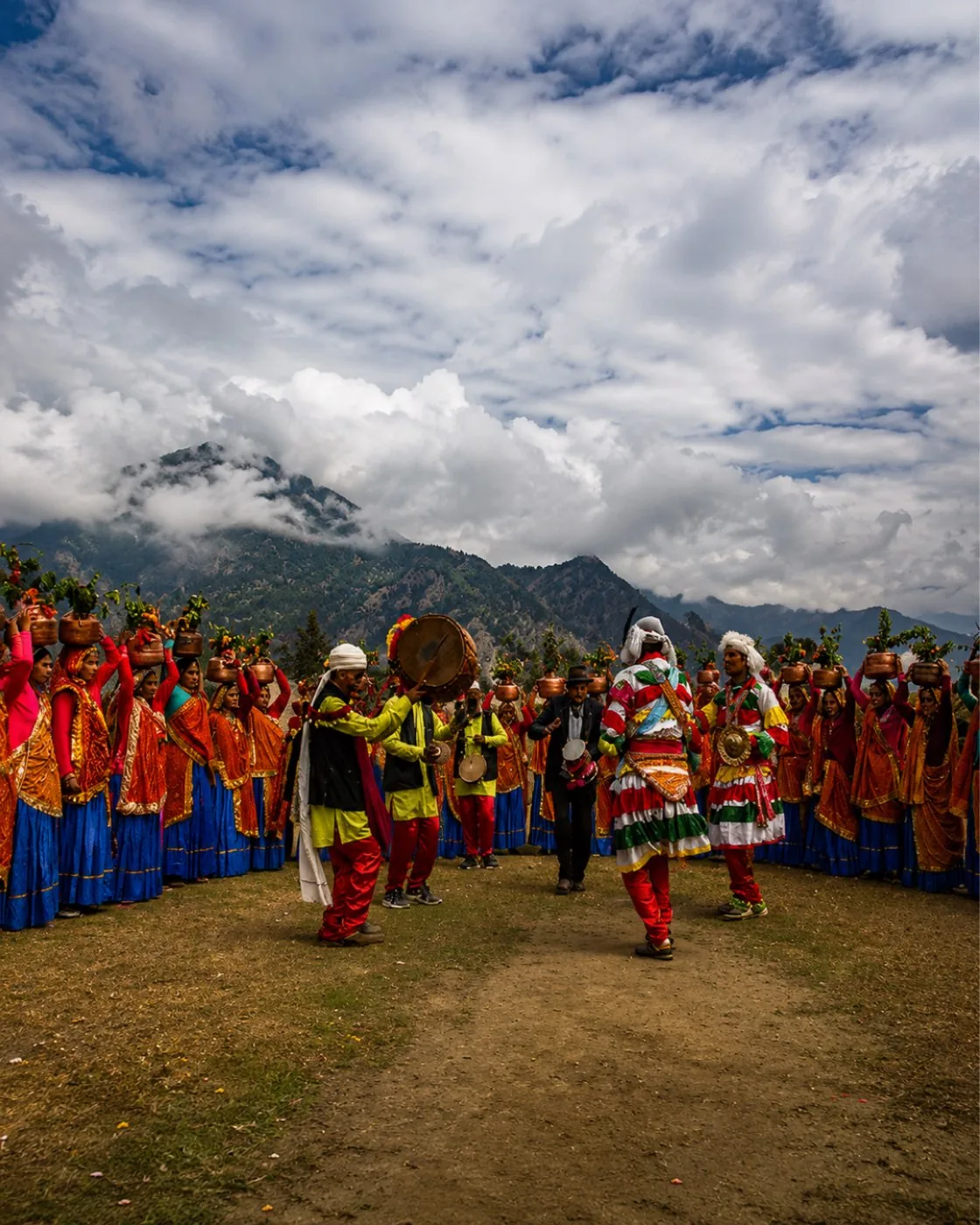 Villagers in bright red and blue carrying copper pots at a hill festival, drummers among them below cloud-covered mountains