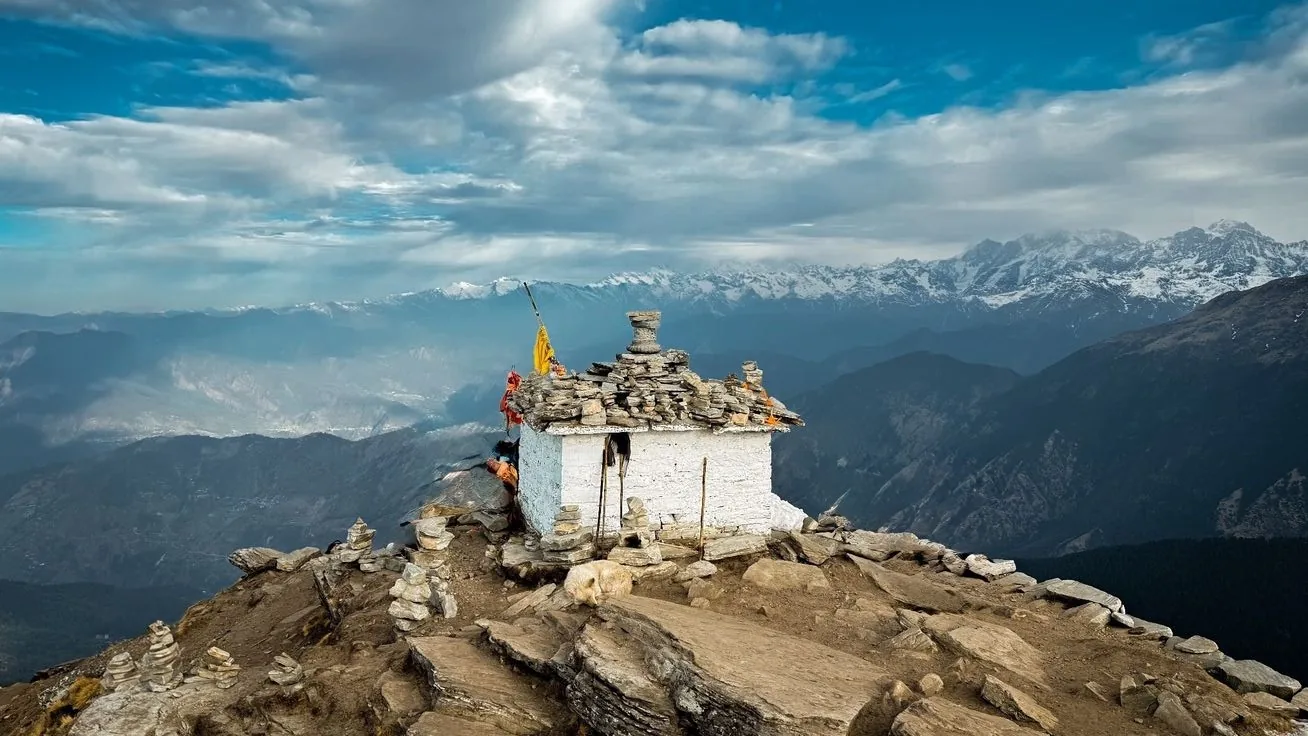 A small whitewashed shrine with a dry-stone slate roof on a rocky summit, votive cairns and a yellow pennant beside it, a long snow-covered range across the horizon and hazy valleys falling away on both sides