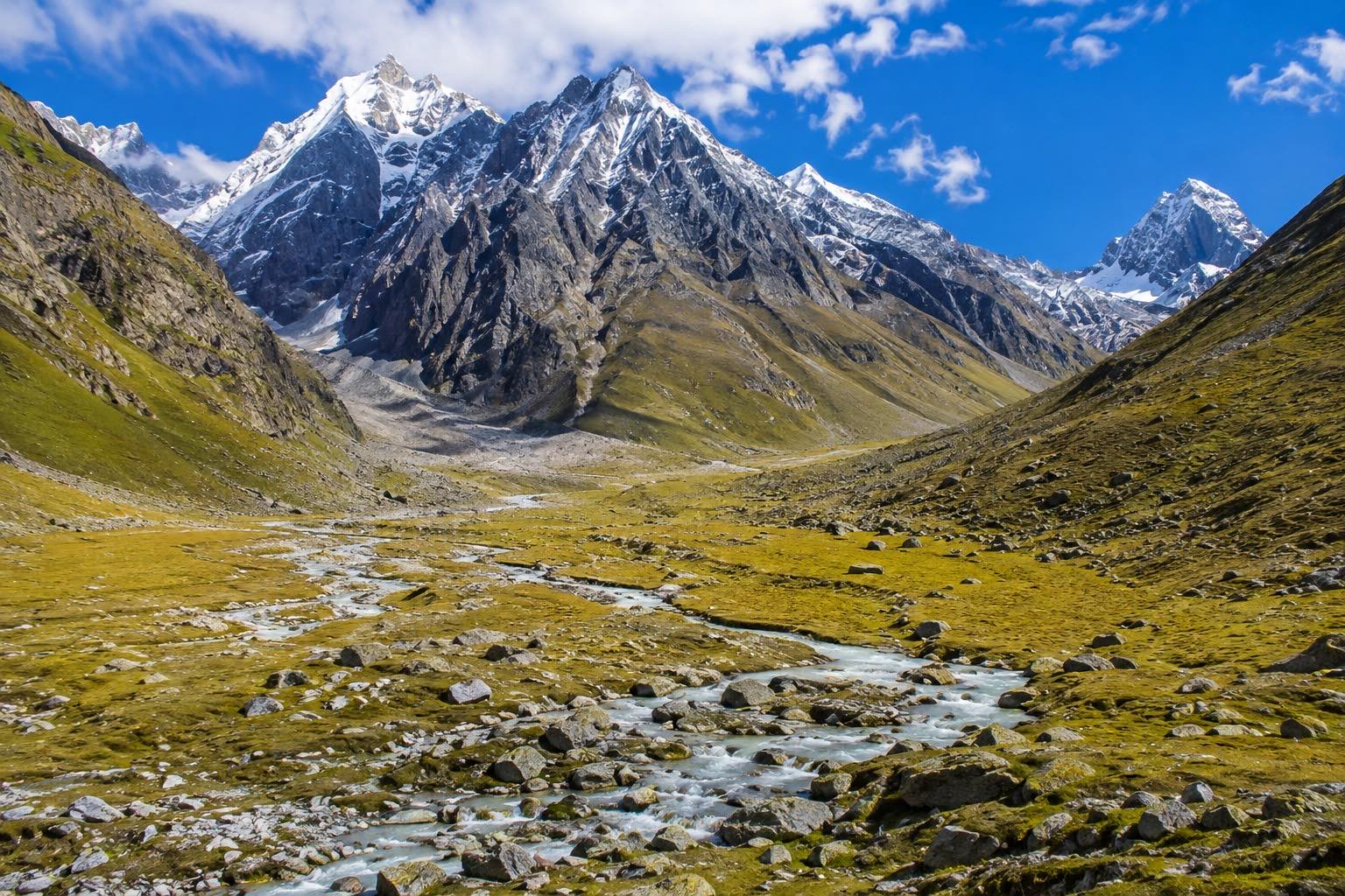 A broad U-shaped valley floor of olive-gold grass with a braided stream threading its stones, a dark rock buttress and snow peaks closing the head