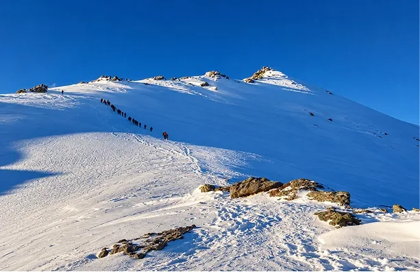 A line of trekkers climbing a broad snow ridge under a deep blue sky