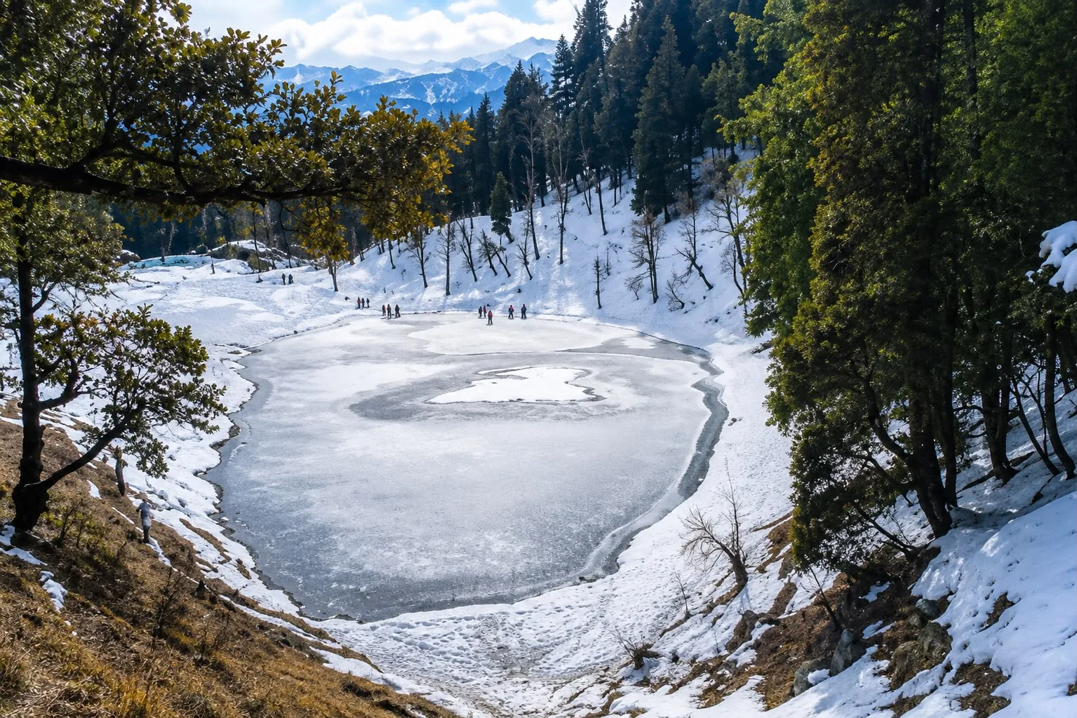 A frozen lake in a wooded bowl with a few figures out on the ice, snow-laden oak and conifer on the slopes above