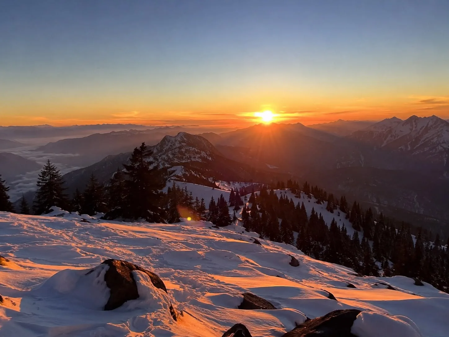 Sunrise over distant ranges above a valley filled with cloud, silhouetted firs and wind-scoured snow in the foreground