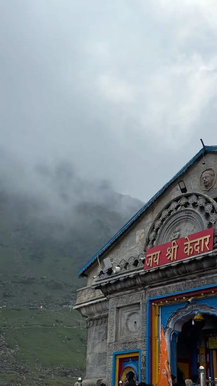 A close view of a carved stone gable with a blue-painted eave and a red board lettered in Devanagari, mist rolling down a green treeless hillside behind
