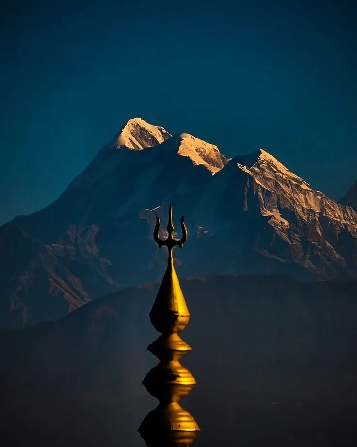 A golden trident finial silhouetted against a deep blue dusk sky, snow peaks behind it still catching the last light