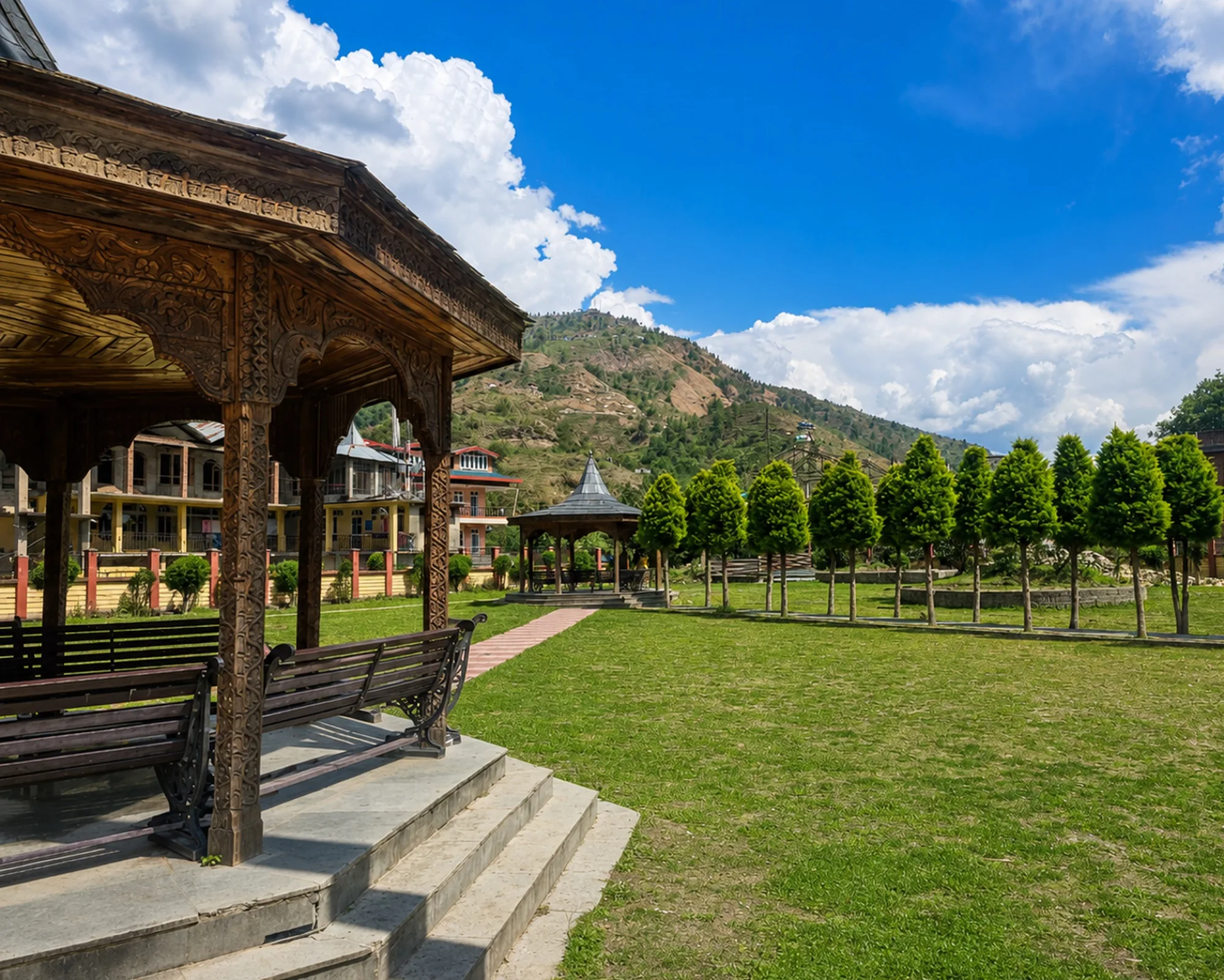 A carved wooden pavilion looking out over a lawn and a line of conifers to a Himalayan hillside