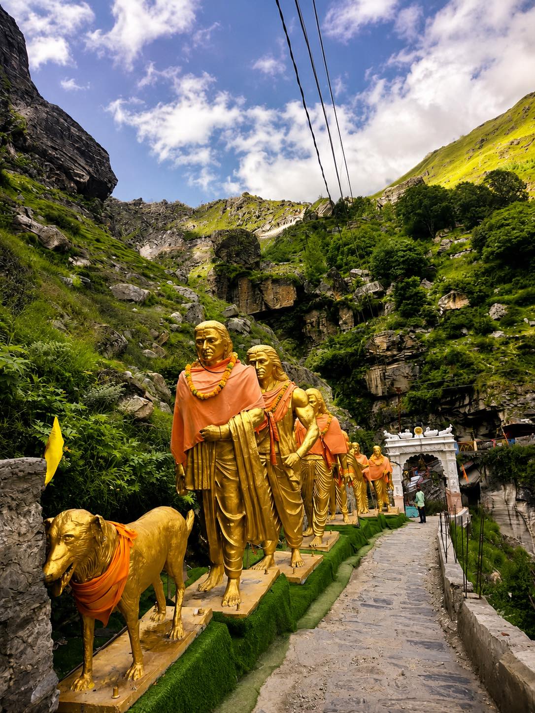 A line of life-size gilded figures draped in orange cloth, a gilded dog at the head of them, on a paved path through a green rocky gorge