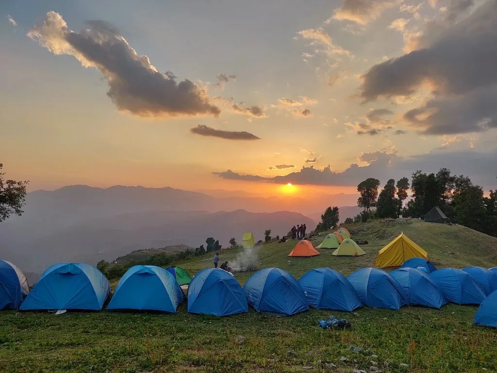 Rows of blue dome tents pitched along a grassy ridge at sunset, a small group standing at the edge beside a fire, oak and deodar to one side and range after range of hazy foothills below