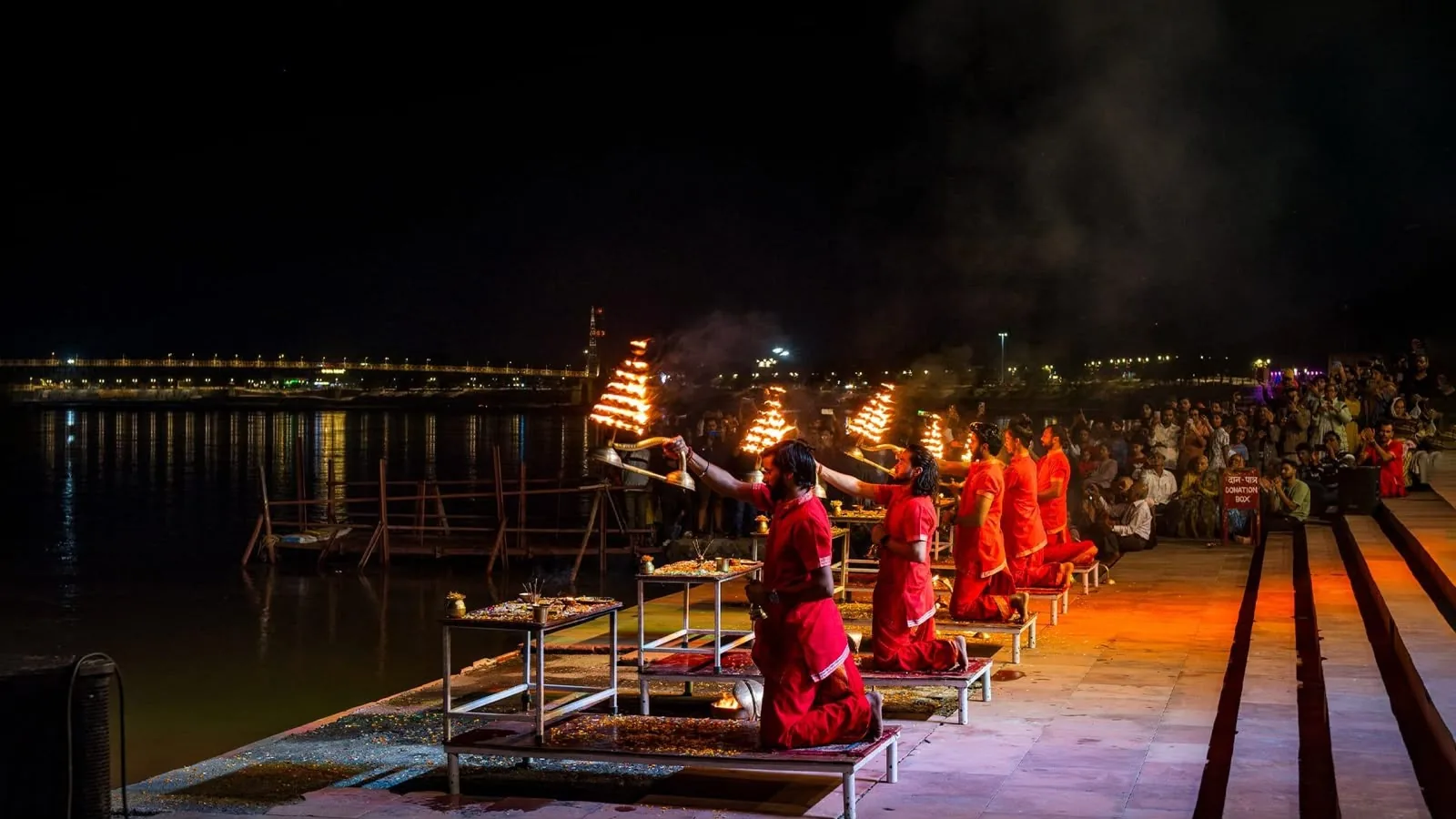 Priests in red holding tiered oil lamps at a river aarti after dark, people watching from the ghat steps
