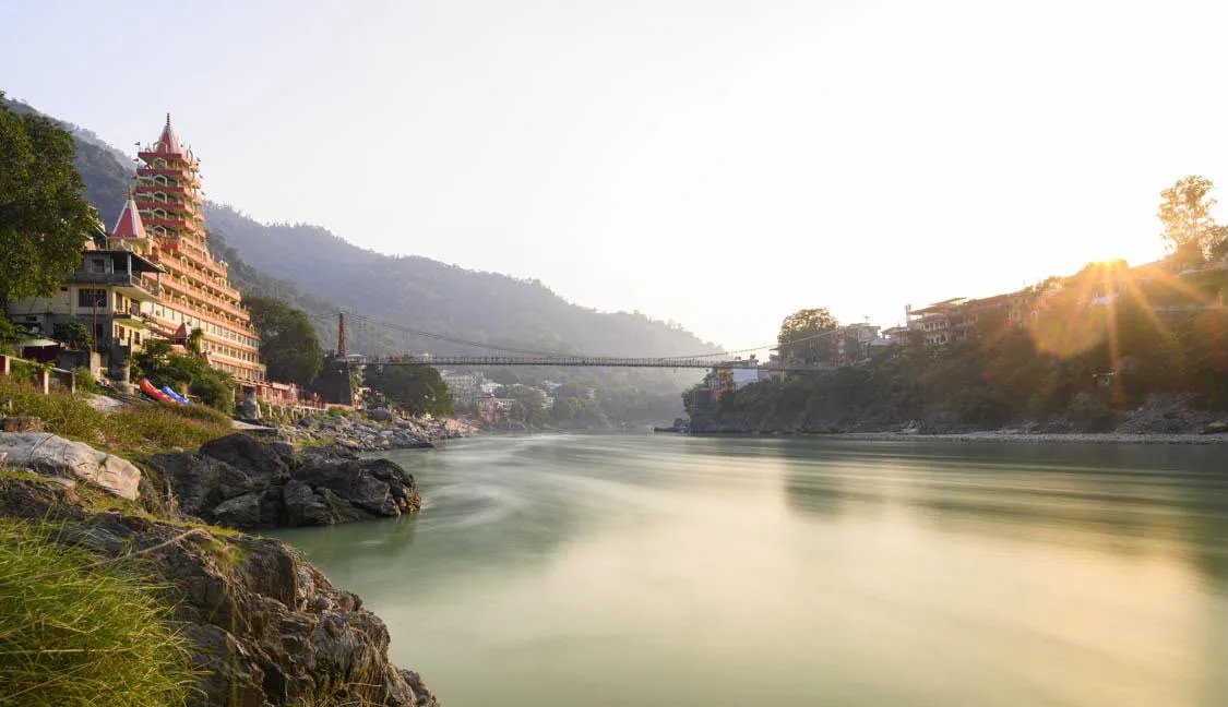 A suspension footbridge across a wide river at sunrise, a tall tiered temple on the near bank and the sun breaking through haze