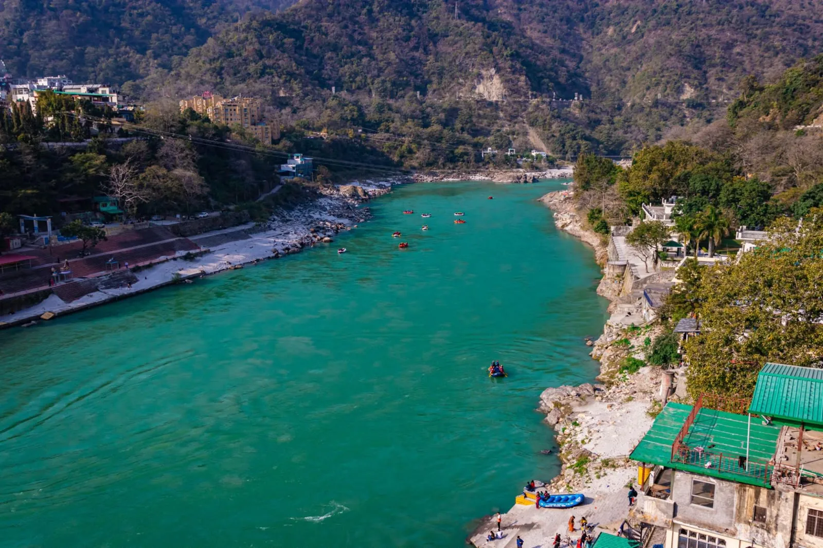 Rafts on a turquoise river seen from above, ghats along one bank and guesthouses on the other