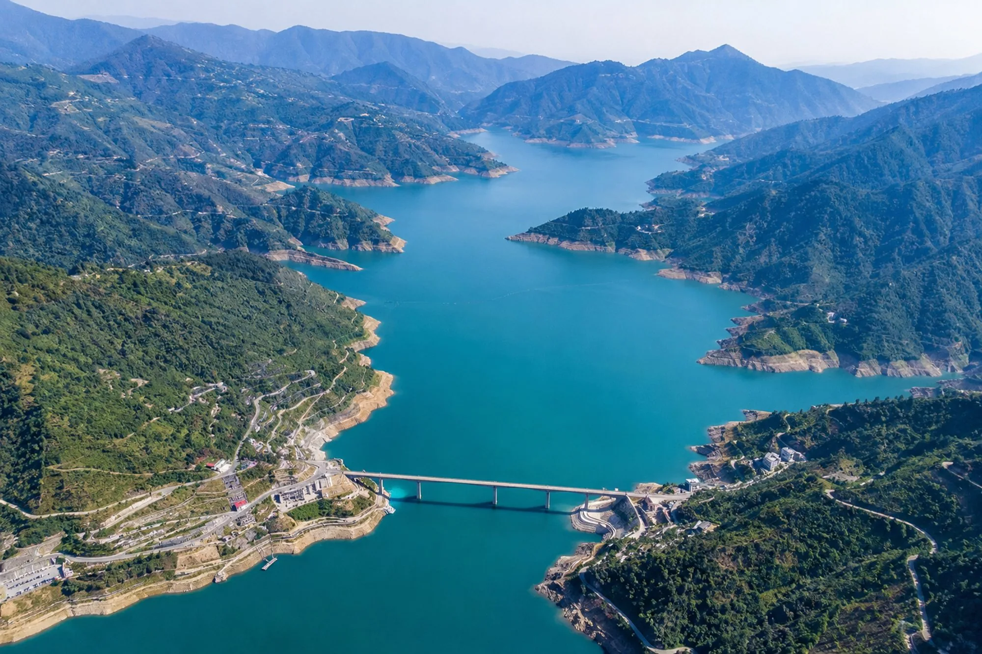 A turquoise reservoir seen from the air, winding between forested ridges with a road bridge crossing it