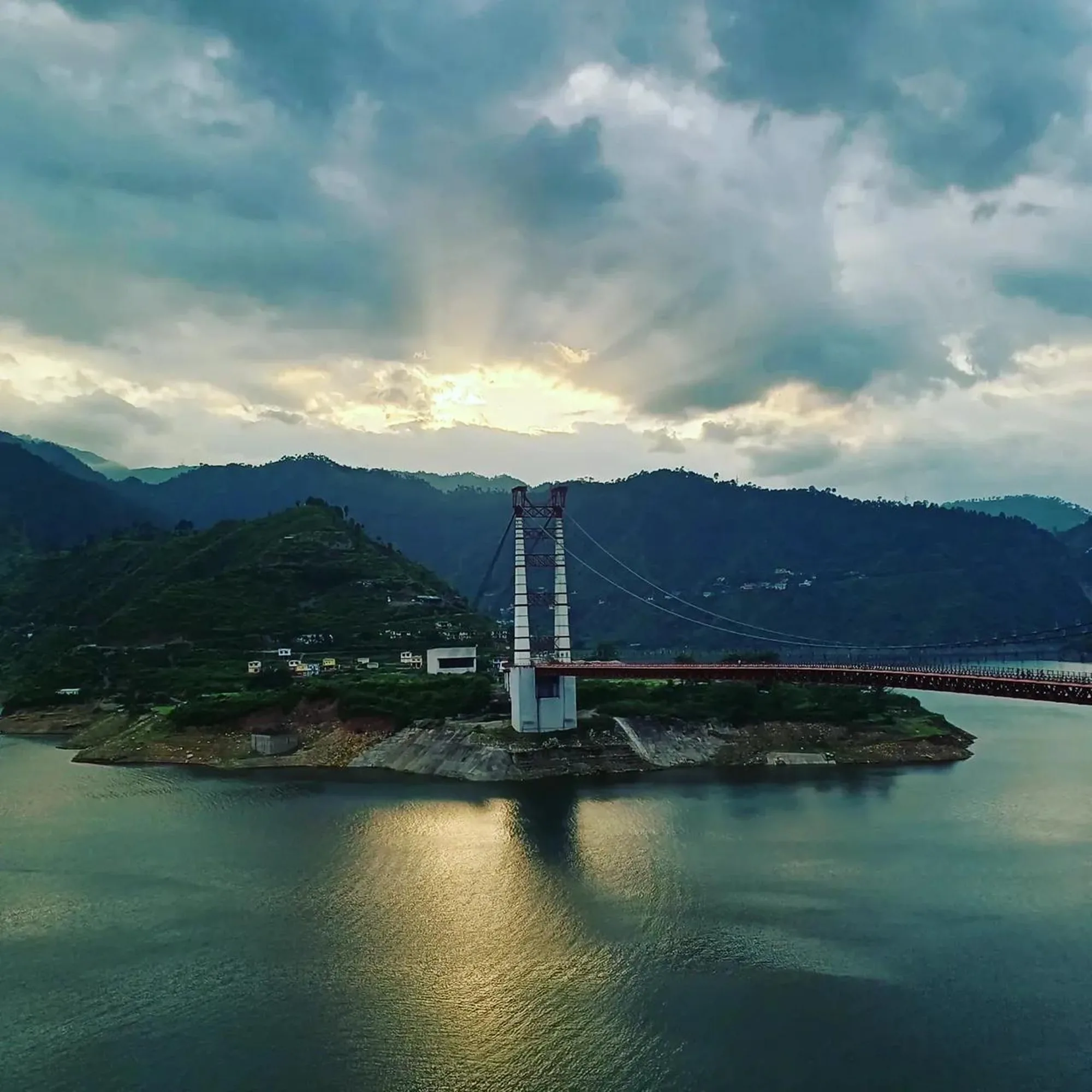 A suspension bridge with a tall white pylon and a red deck crossing a reservoir, under broken cloud at sunset