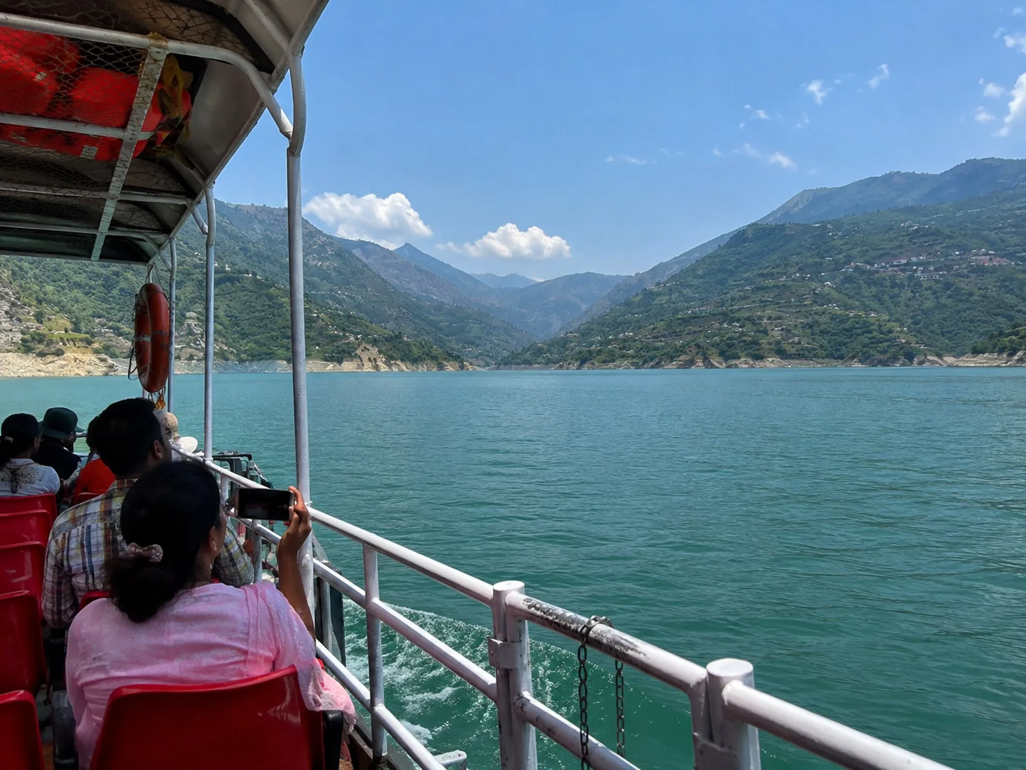 Passengers on the open deck of a ferry crossing a reservoir, with terraced hillsides rising on both sides