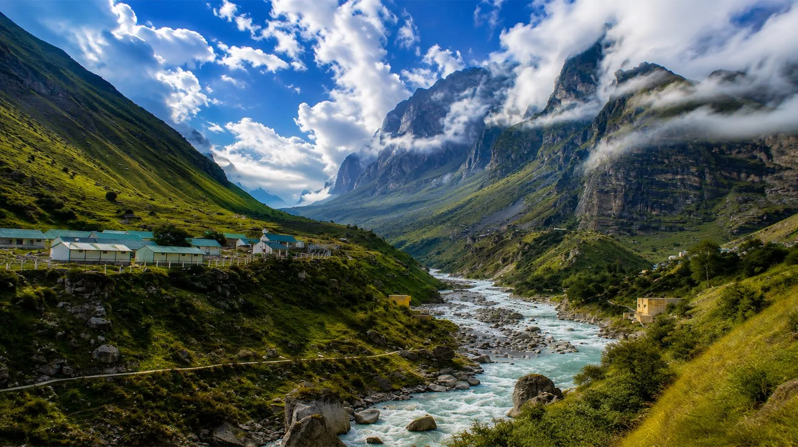 A glacial river running through a green Himalayan valley, with a cluster of huts below cloud-covered cliffs.