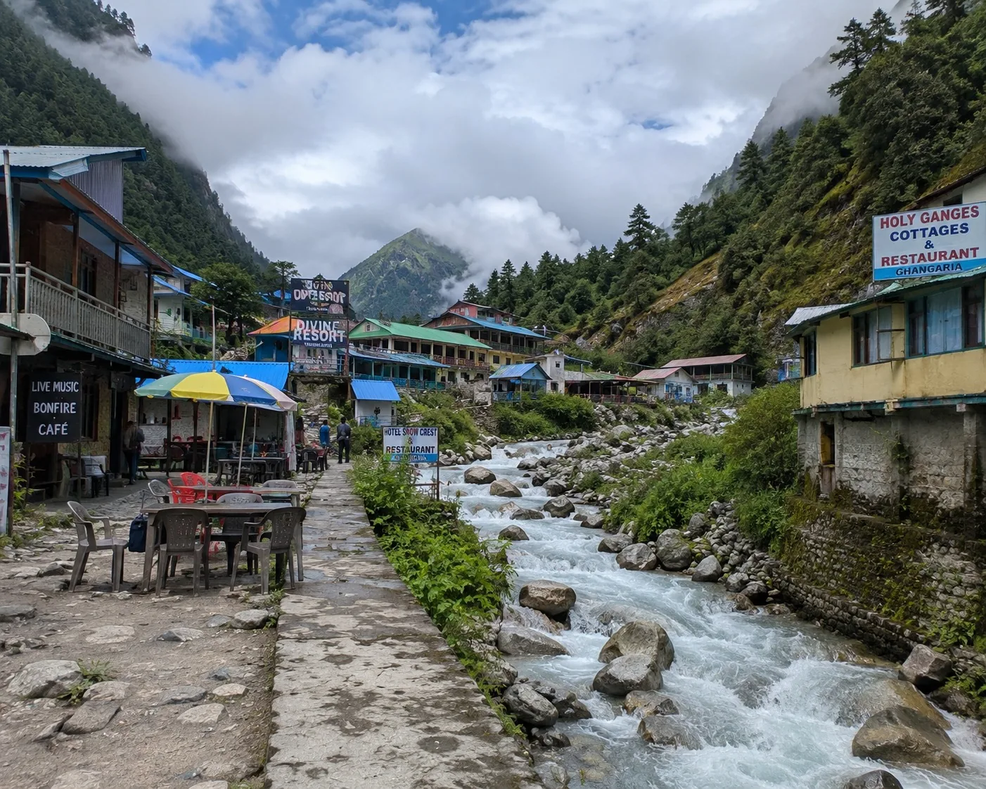A village street of tin-roofed lodges and cafe tables beside a boulder-strewn glacial torrent, conifer slopes and cloud above