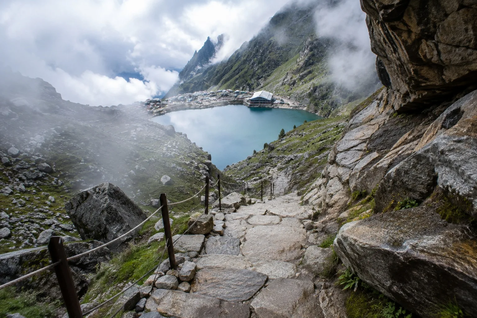 A stone stepped path with rope railings descending to a green-blue mountain tarn, a large sloped-roof building and a cluster of huts on the far shore, mist across the rock walls