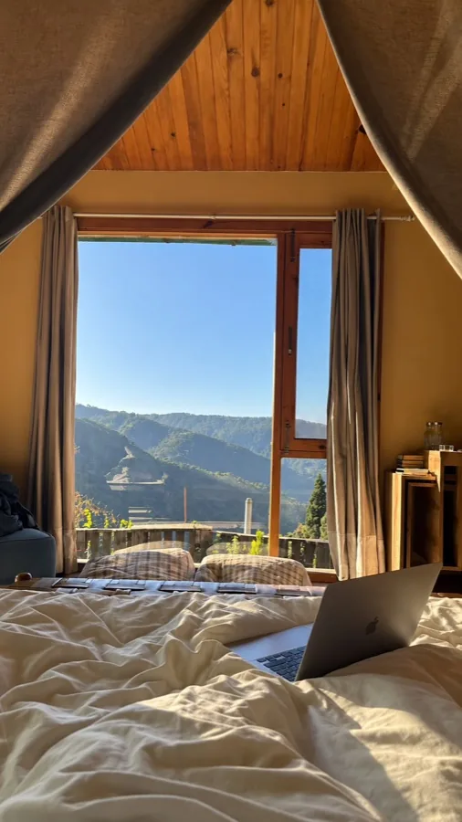 Looking out from a bed in a wooden cabin through a tall window to forested Himalayan ridges, a laptop open on the covers