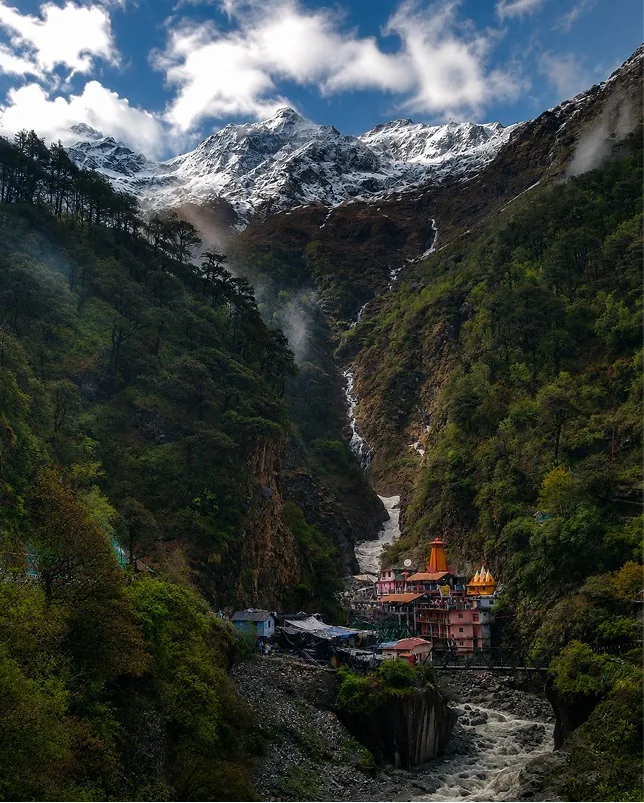 A temple in a forested gorge below a waterfall and snow-covered peaks
