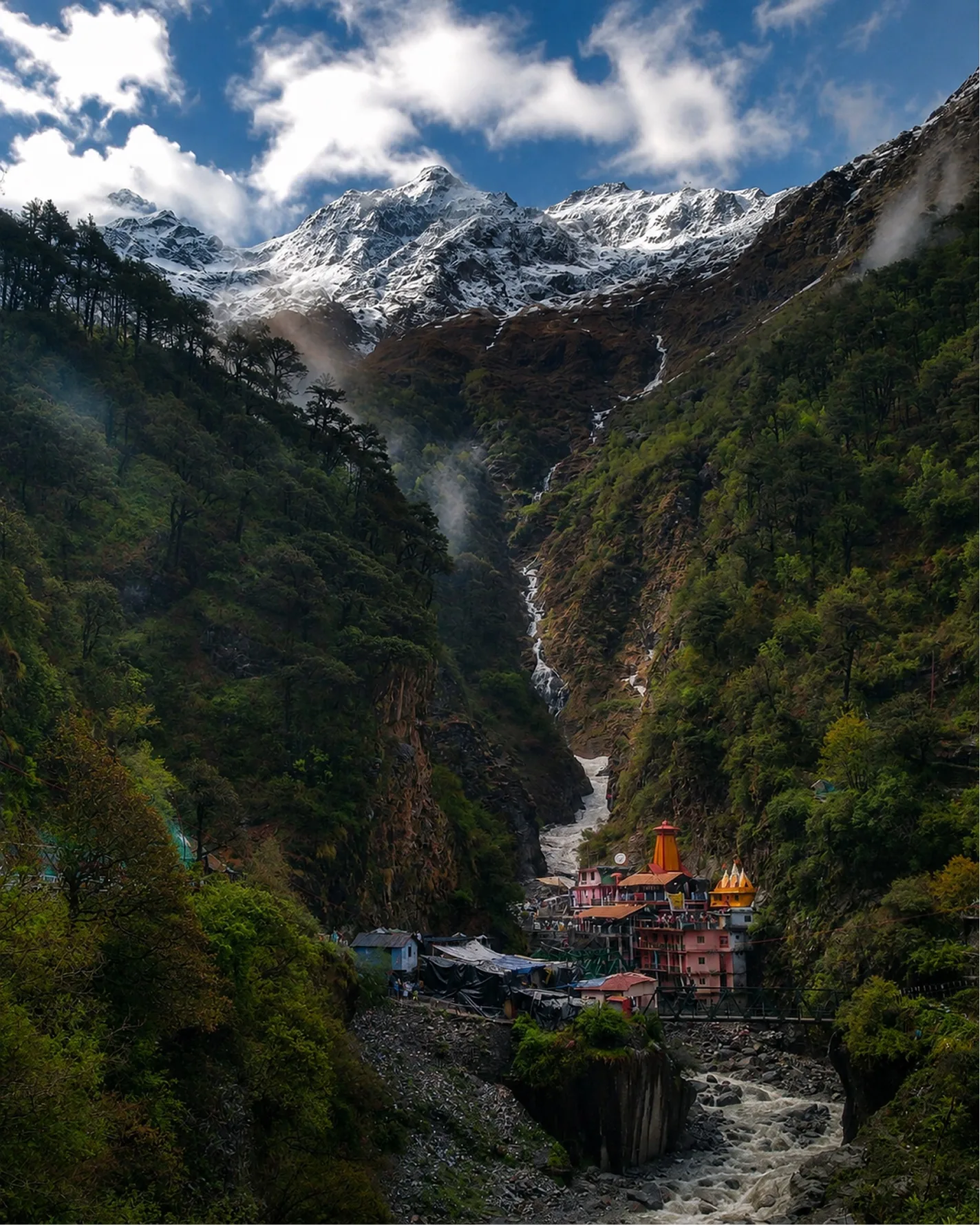 A small temple settlement in a forested gorge below snow peaks and a waterfall