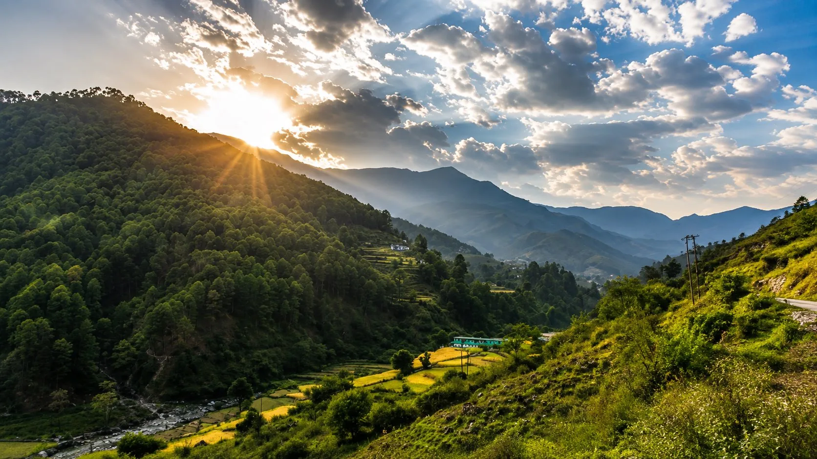 Sun breaking behind a pine ridge over a terraced valley of cut fields and scattered houses, under a cloud-broken sky