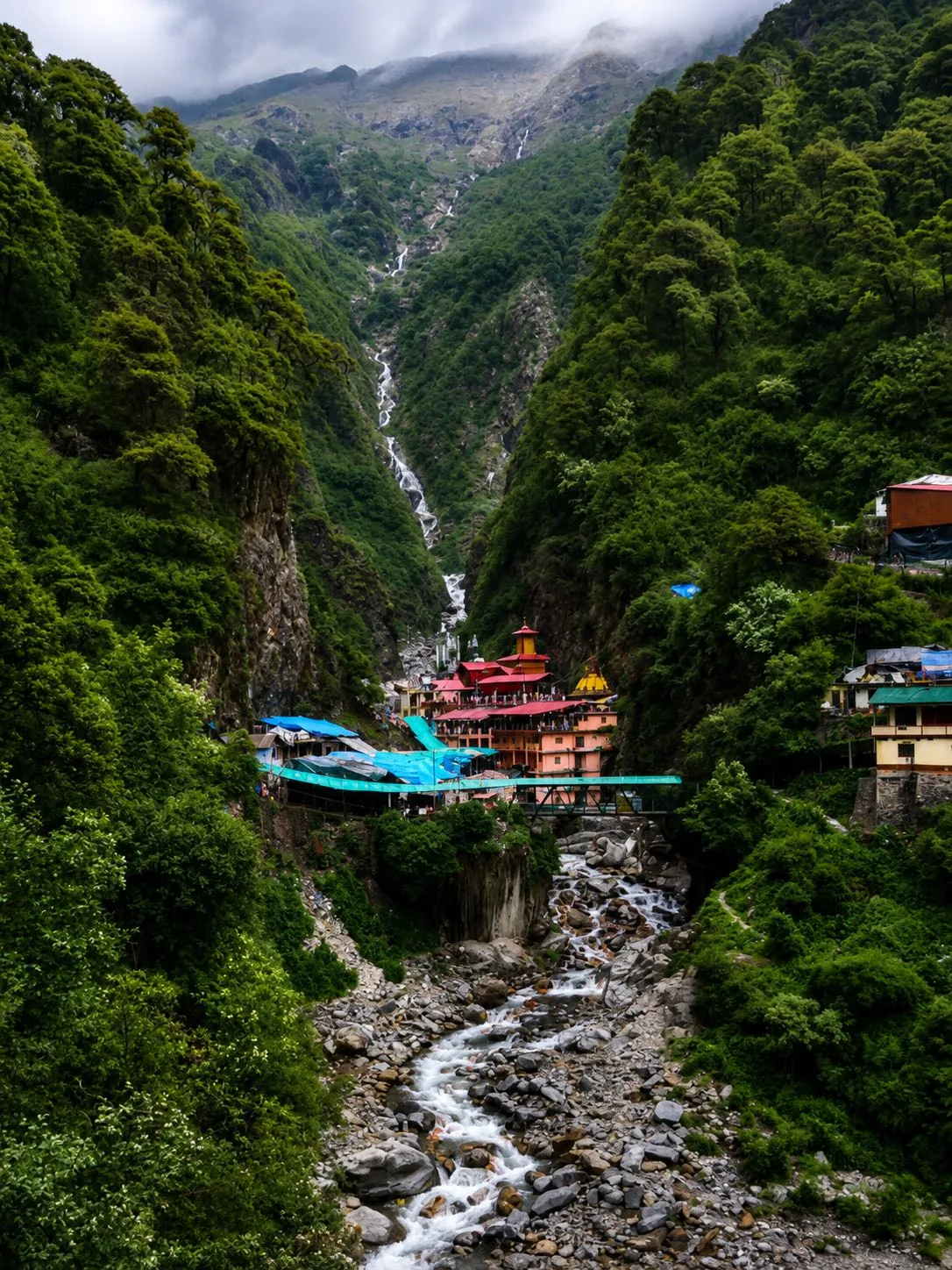 A temple complex with red and gold roofs deep in a steep forested gorge, waterfalls threading the cliffs above it and a boulder torrent below