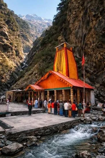 A small orange and yellow shrine on a paved riverside terrace at the foot of a cliff, a queue of pilgrims at its door and a snow peak beyond