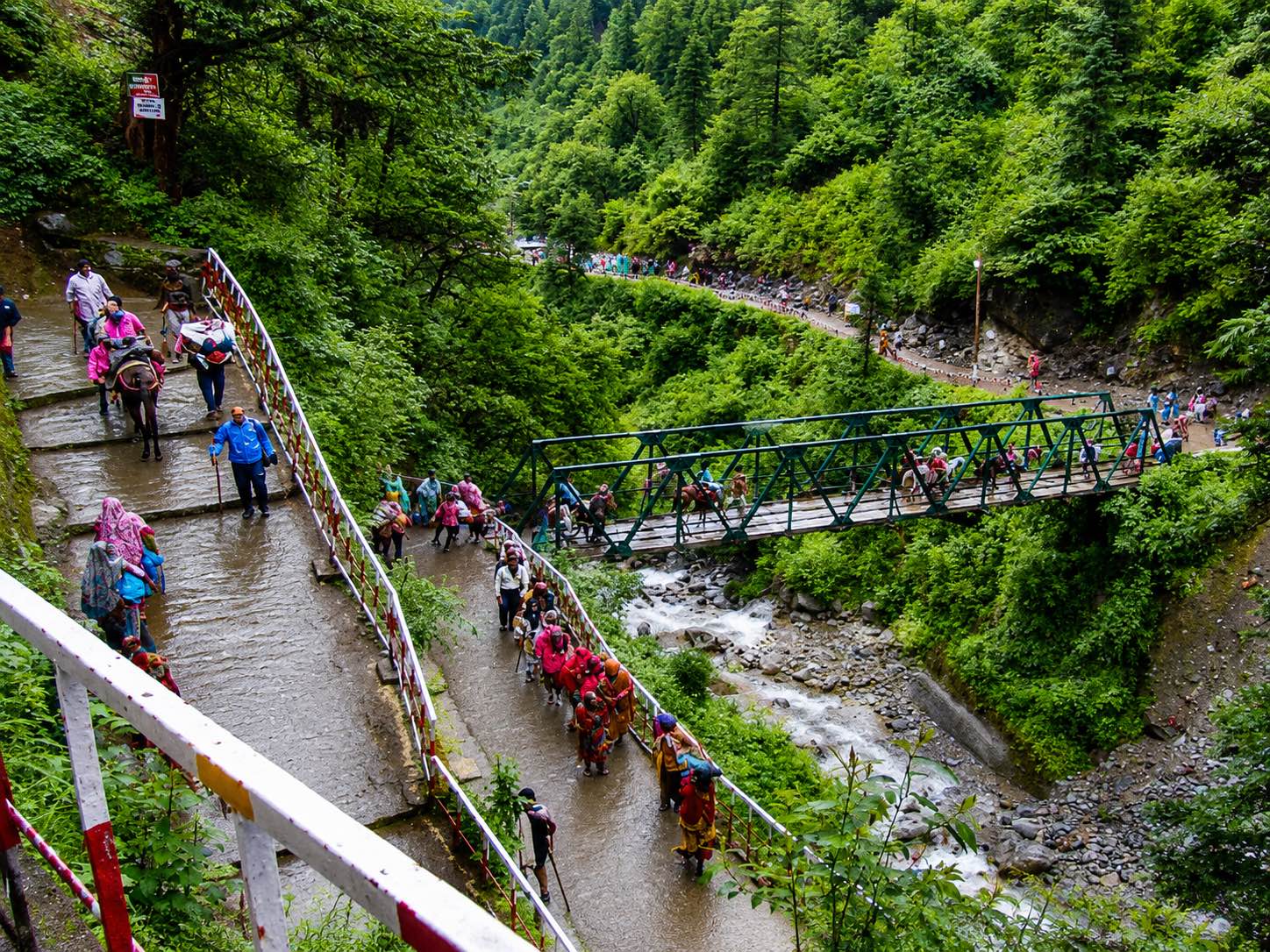 A wet paved switchback trail through conifer forest with pilgrims in bright rain gear and pack ponies climbing, and a green truss bridge over the stream